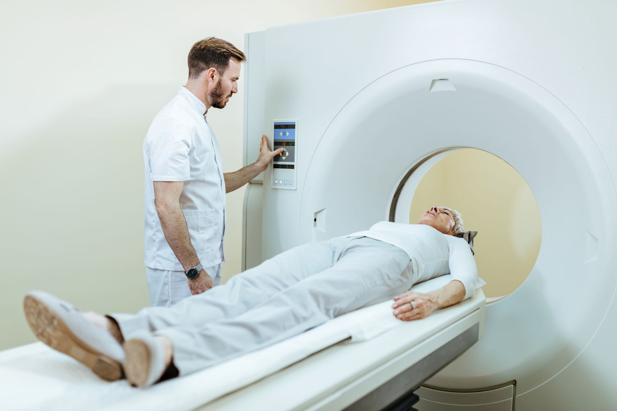 Man clinician standing next to an MRI machine, whilst a lady lays on the bed. The man is pushing a button on the outside of the machine.