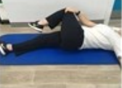 A person lying on their back on an exercise mat, with the right leg out straight and the left leg bent and crossed over the body. Their right hand is holding the knee of their left leg.