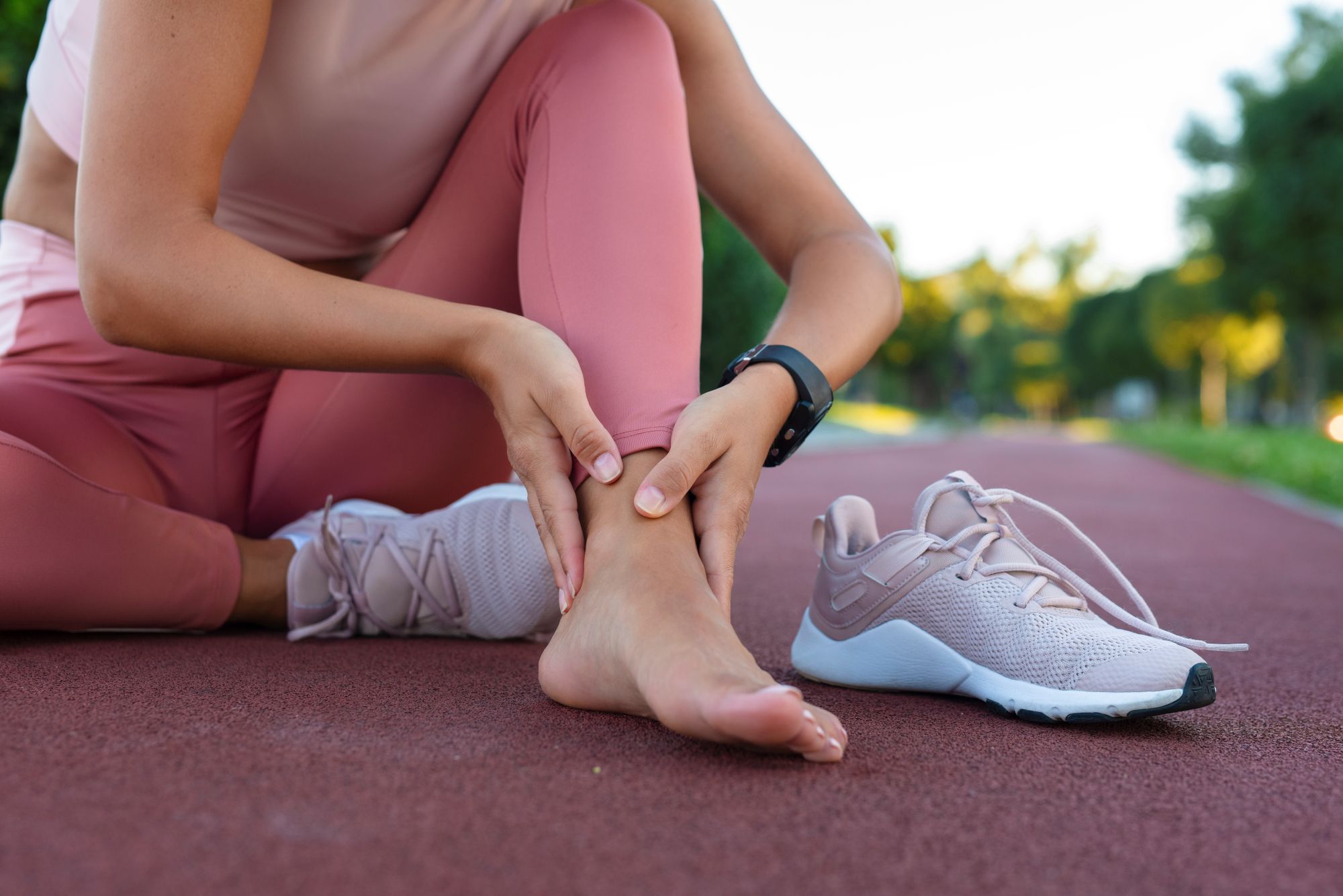 Woman dressed in exercise clothes, sitting on a running track with her barefoot resting on the ground and holding her ankle in pain. Her trainer sits besides her.