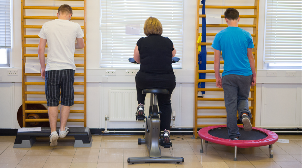 A photograph of a gym based rehabilitation session with 3 patients with their backs to the camera. one man step sup onto a step, next to an  elderly woman on an exercise bicycle and finally a young man jumping on a trampette, all in a row.. 