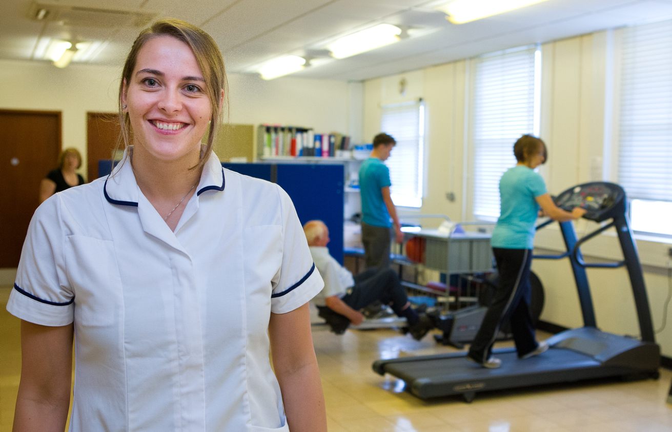 Woman clinician in white uniform standing in the forefront smiling at the camera. Behind her is a woman on a treadmill, and elderly man on a rowing machine and a second man standing up with his back to the camera.