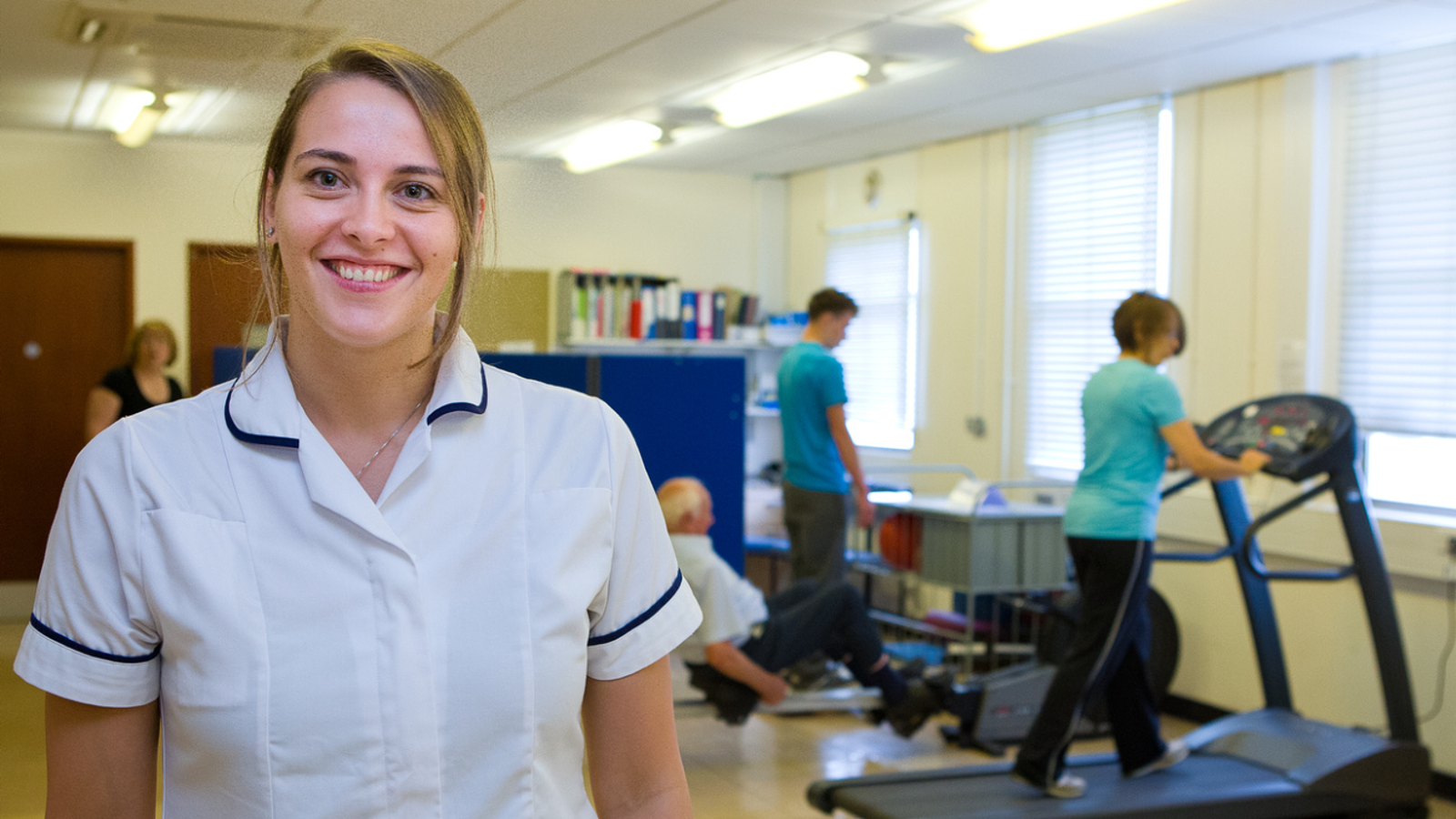 Woman clinician in white uniform standing in the forefront smiling at the camera. Behind her is a woman on a treadmill, and elderly man on a rowing machine and a second man standing up with his back to the camera.