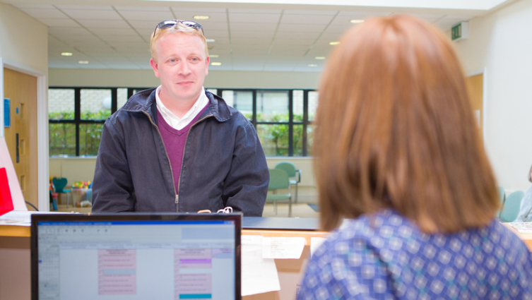 White male approaches a clinic reception desk with the back of a female receptionists head, a computer screen and paperwork are placed.